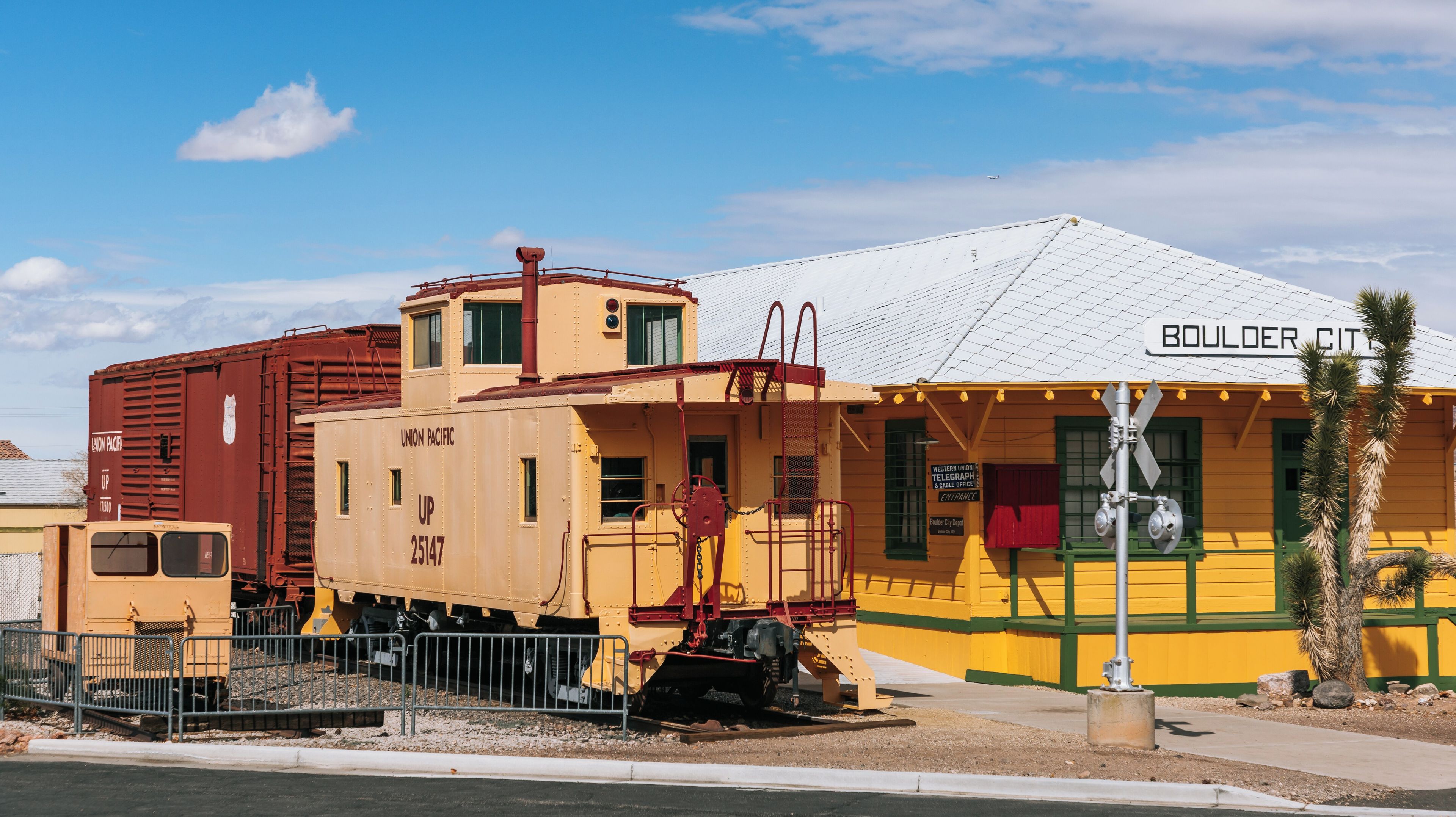 A Union Pacific train car, a yellow building with 'BOULDER CITY' signage, and a clear blue sky.
