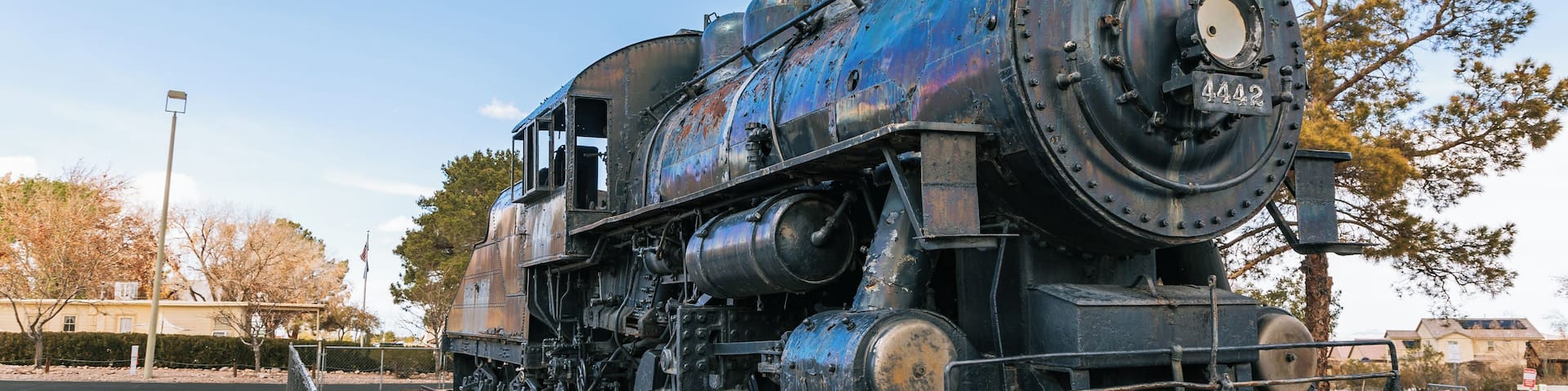 A vintage steam locomotive with visible wear and rust, stationed on tracks with a clear sky and trees in the background.