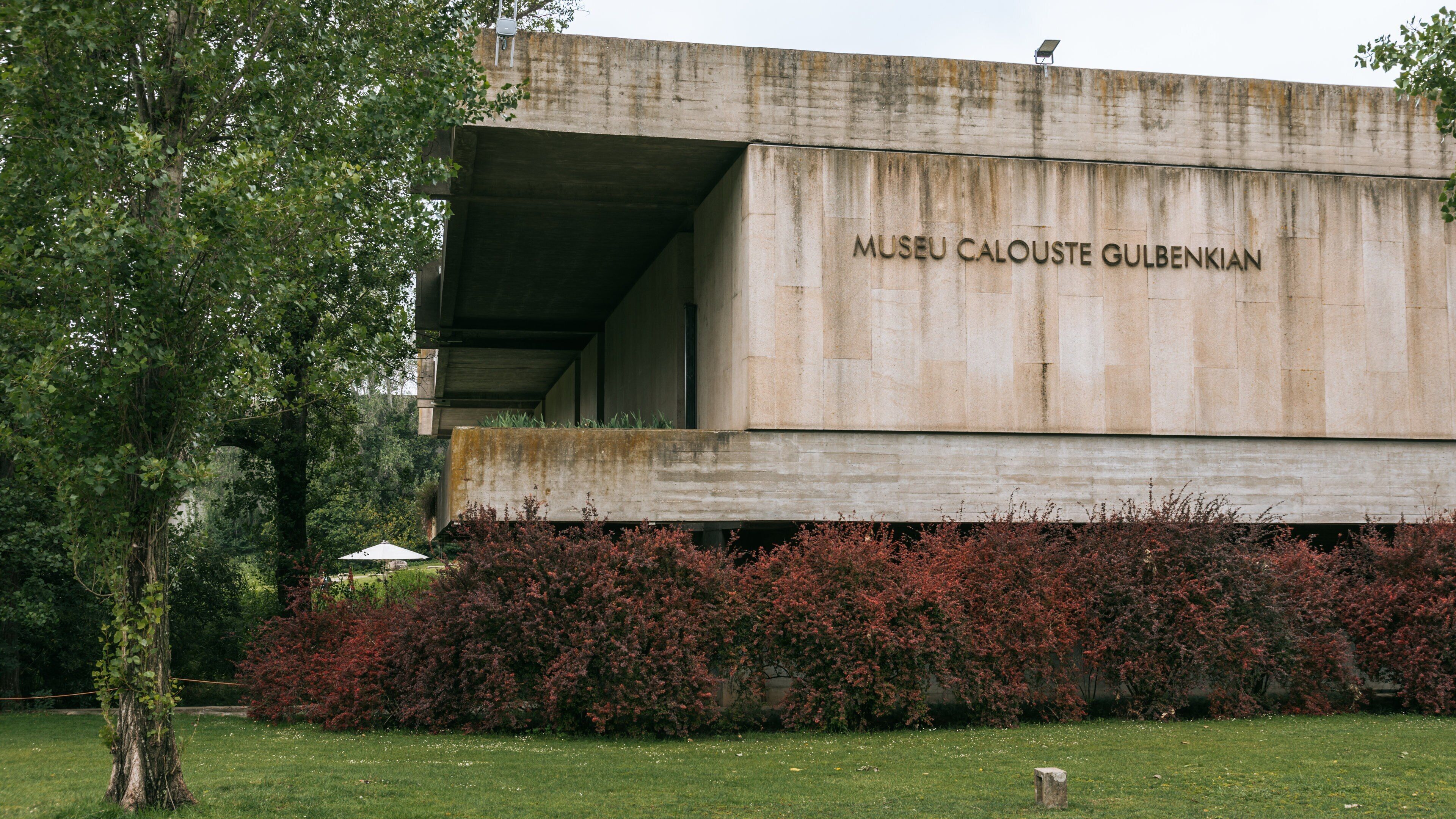A concrete building with the inscription "MUSEU CALOUSTE GULBENKIAN" and a surrounding garden with red foliage.
