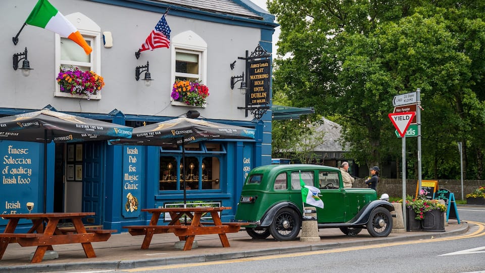 A pub with a blue exterior, American and Irish flags, and a vintage green car parked outside.
