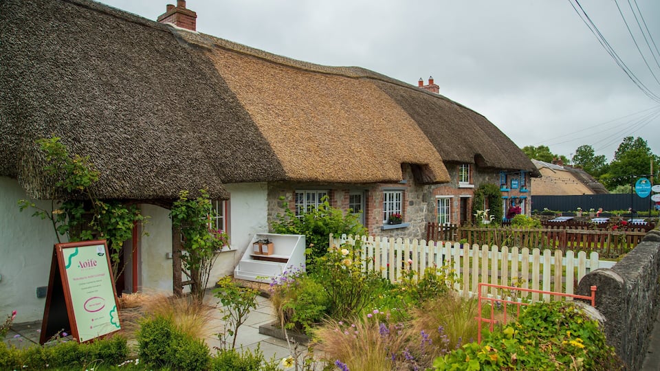 A thatched-roof cottage with a white picket fence and a signboard.
