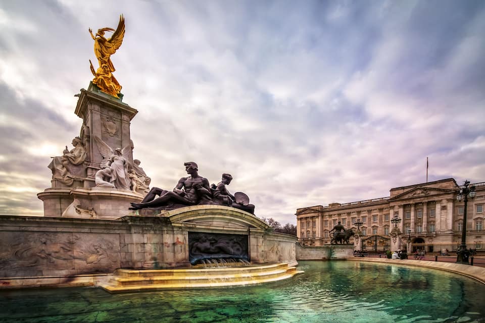 Ein klassischer Brunnen mit Statuen, ein goldener Engel und ein neoklassizistisches Gebäude im Hintergrund.