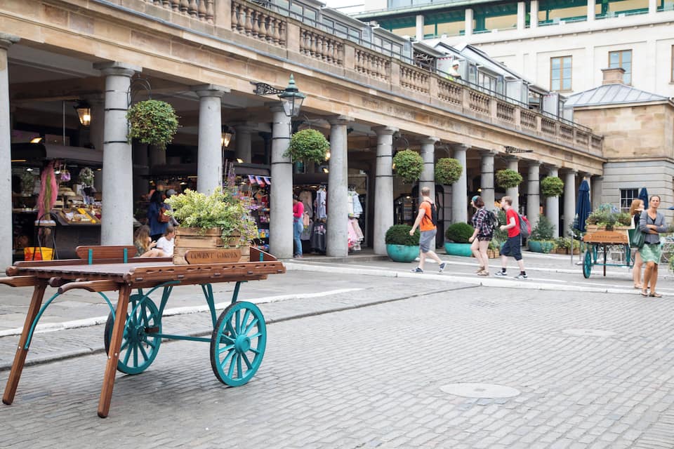 A cobblestone street with a wooden cart and a cart with a sign reading "COVENT GARDEN."