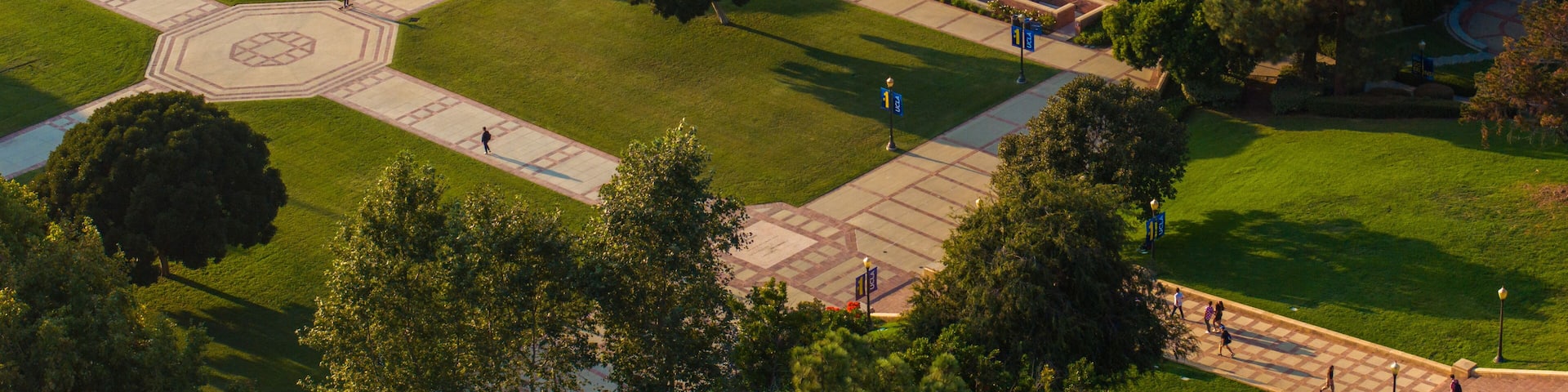 Aerial view of a grand university campus with red-bricked buildings, green lawns, and classical architecture, featuring a sports field, pond, and serene outdoor seating areas.