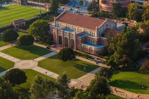 Aerial view of a grand university campus with red-bricked buildings, green lawns, and classical architecture, featuring a sports field, pond, and serene outdoor seating areas.