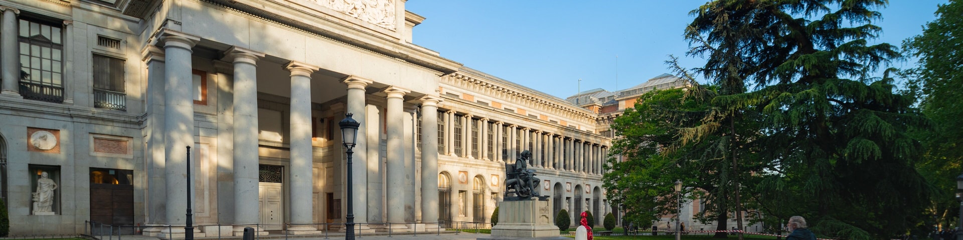 Um edifício neoclássico com colunas, uma bandeira e uma estátua em uma praça.