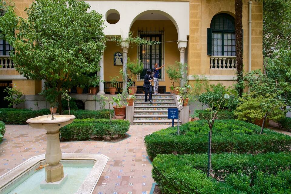 A courtyard with a fountain, potted plants, and a building with arched windows and a balcony.