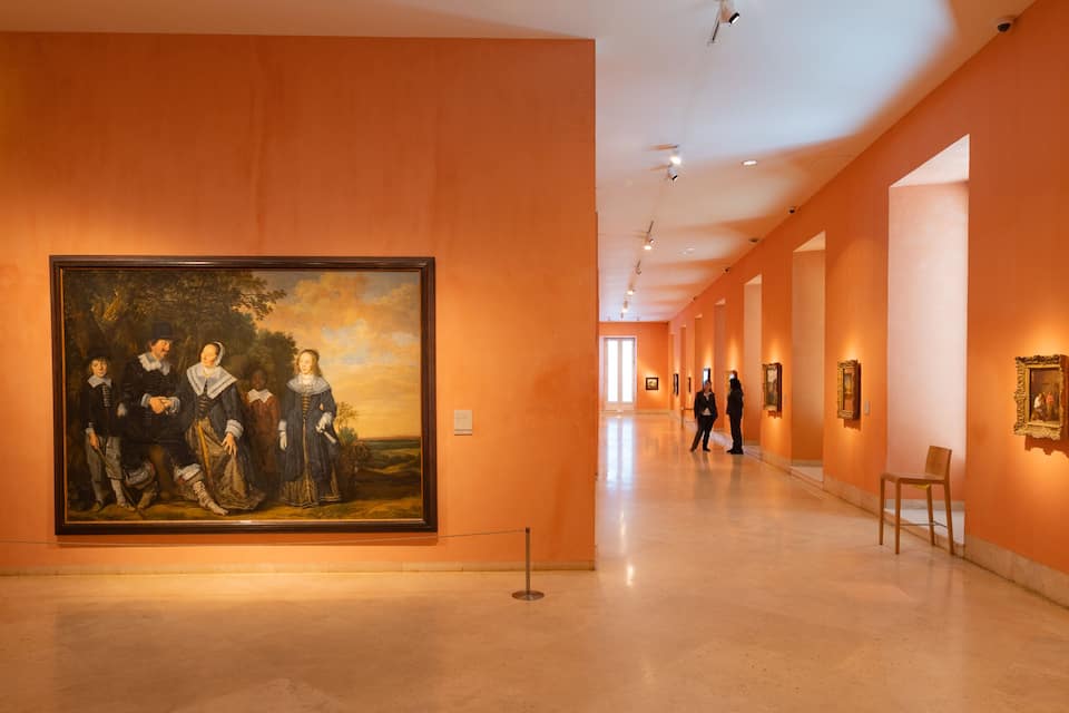 Una galería de arte con un gran cuadro clásico en la pared izquierda y varias obras de arte enmarcadas en la derecha.