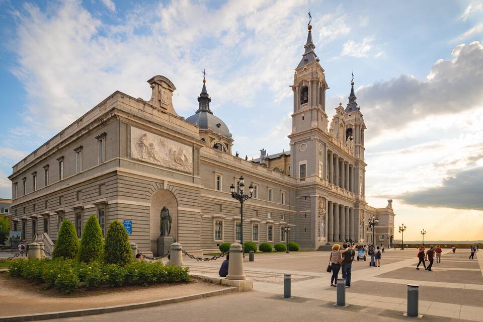 Un bâtiment historique à l’architecture classique, avec des colonnes et un dôme, entouré d’une place animée où se côtoient les gens et la verdure.