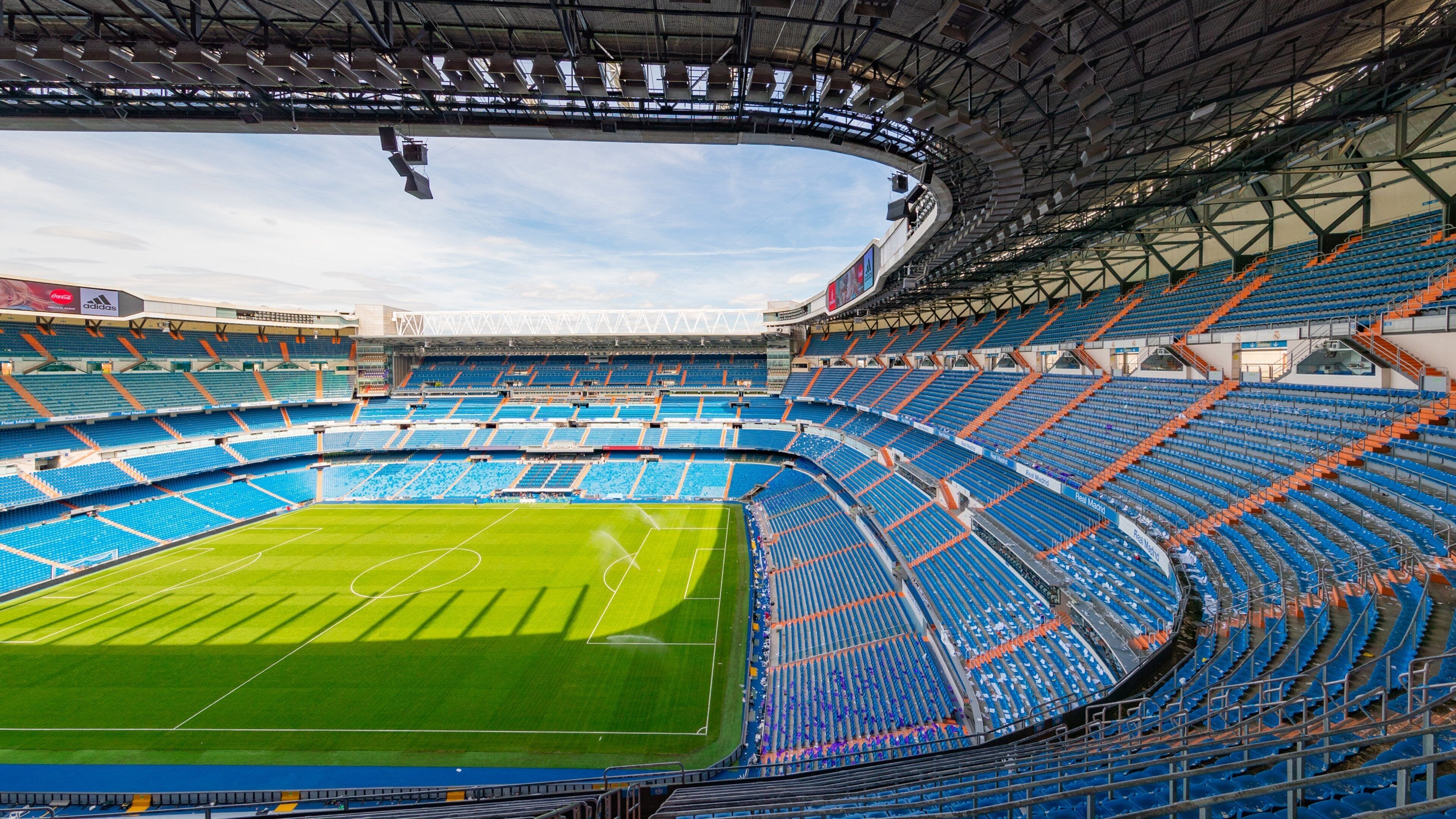 Un estadio amplio y vacío con butacas azules y anaranjadas, un campo verde y un techo alto con luminarias.