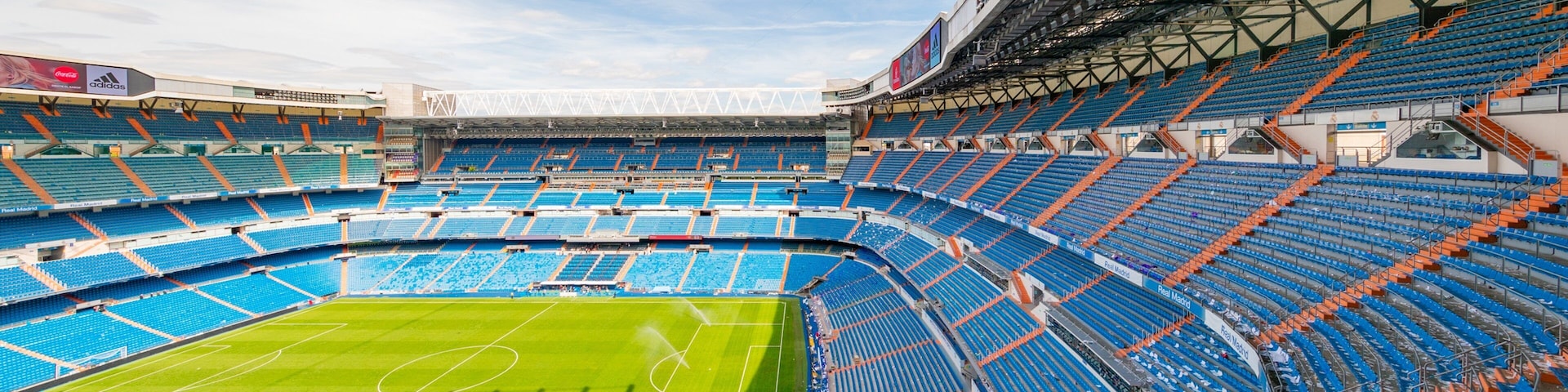 Un estadio amplio y vacío con butacas azules y anaranjadas, un campo verde y un techo alto con luminarias.