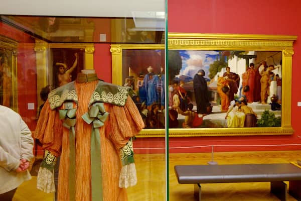 Une exposition dans un musée présentant un mannequin vêtu d’un costume traditionnel et deux grandes peintures accrochées au mur.