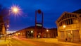 Une rue au crépuscule, avec une enseigne « MOSI » bien visible, des lampadaires et un bâtiment historique doté de fenêtres en arc de cercle.