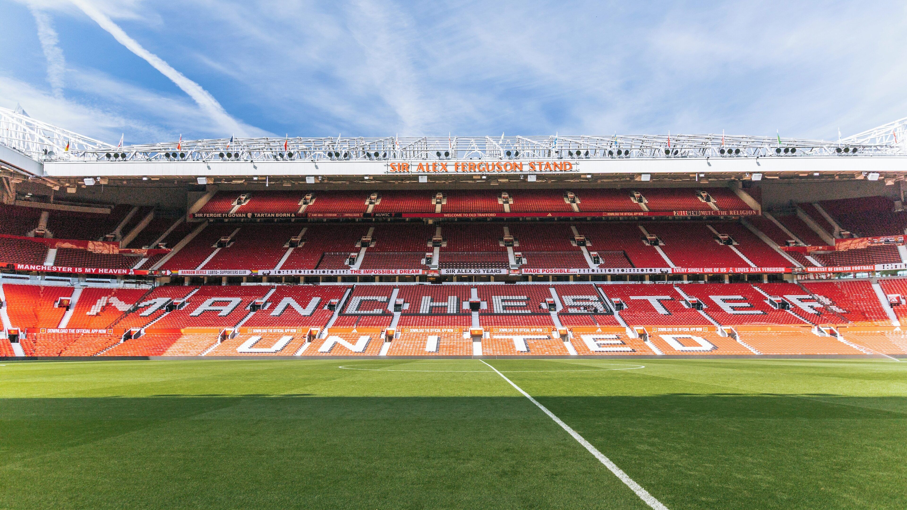 Old Trafford stadium in Manchester shows vibrant red seating and lush green pitch beneath a clear blue sky