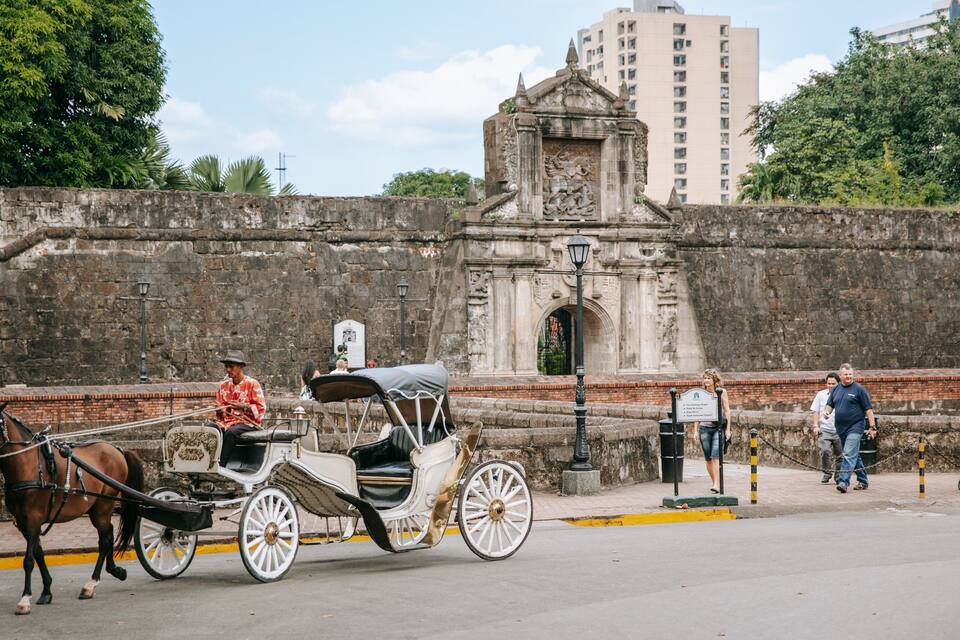 Uma carruagem puxada por cavalos numa rua, com um muro de pedra histórico e um portão ao fundo.