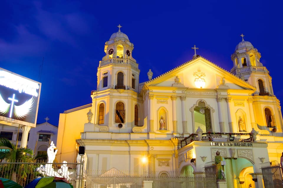 Uma igreja bem iluminada, com duas torres de sino e uma grande cruz na fachada.