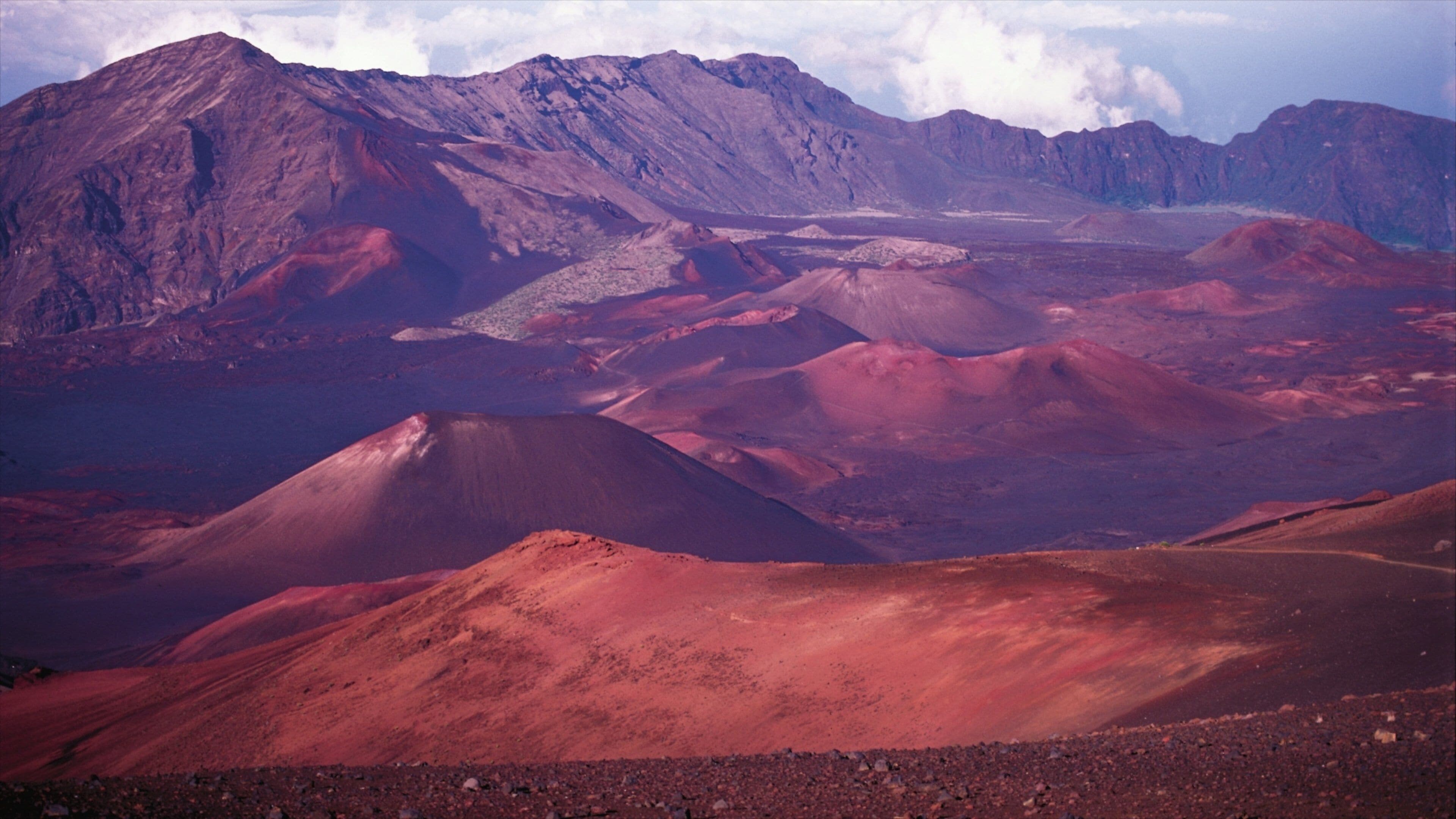 A vast volcanic landscape with prominent red-hued mountains and a distant valley.