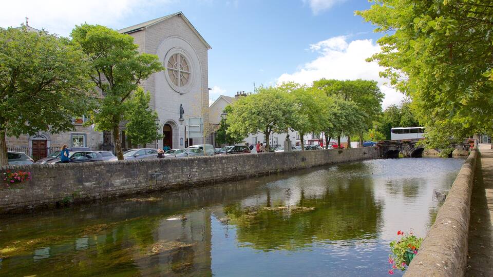A canal with a stone wall, a church with a circular window, and parked cars along the side.