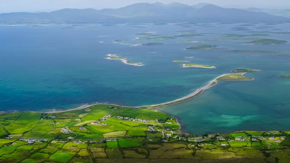 An aerial view of a coastal area with green fields, a winding road, and a large body of water with clear blue hues.