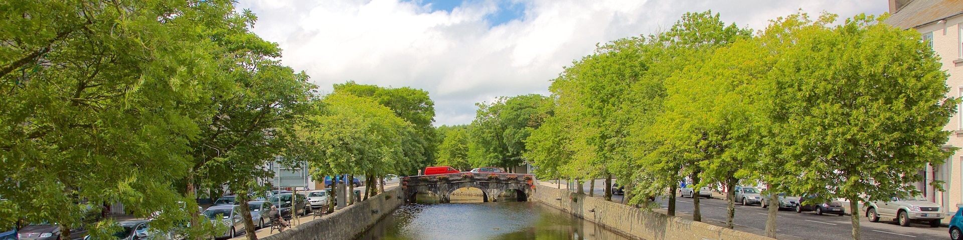 A canal with a stone bridge, lined with trees and parked cars.