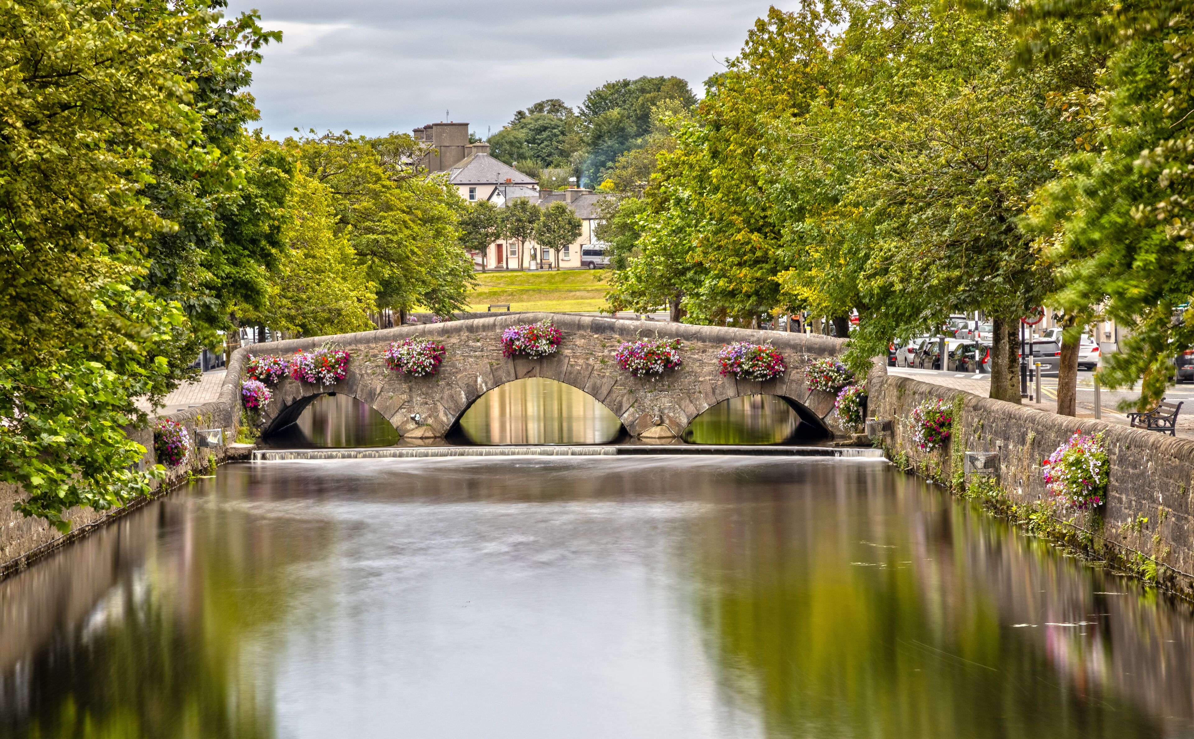 A stone bridge with flower boxes over a calm river, surrounded by lush green trees.