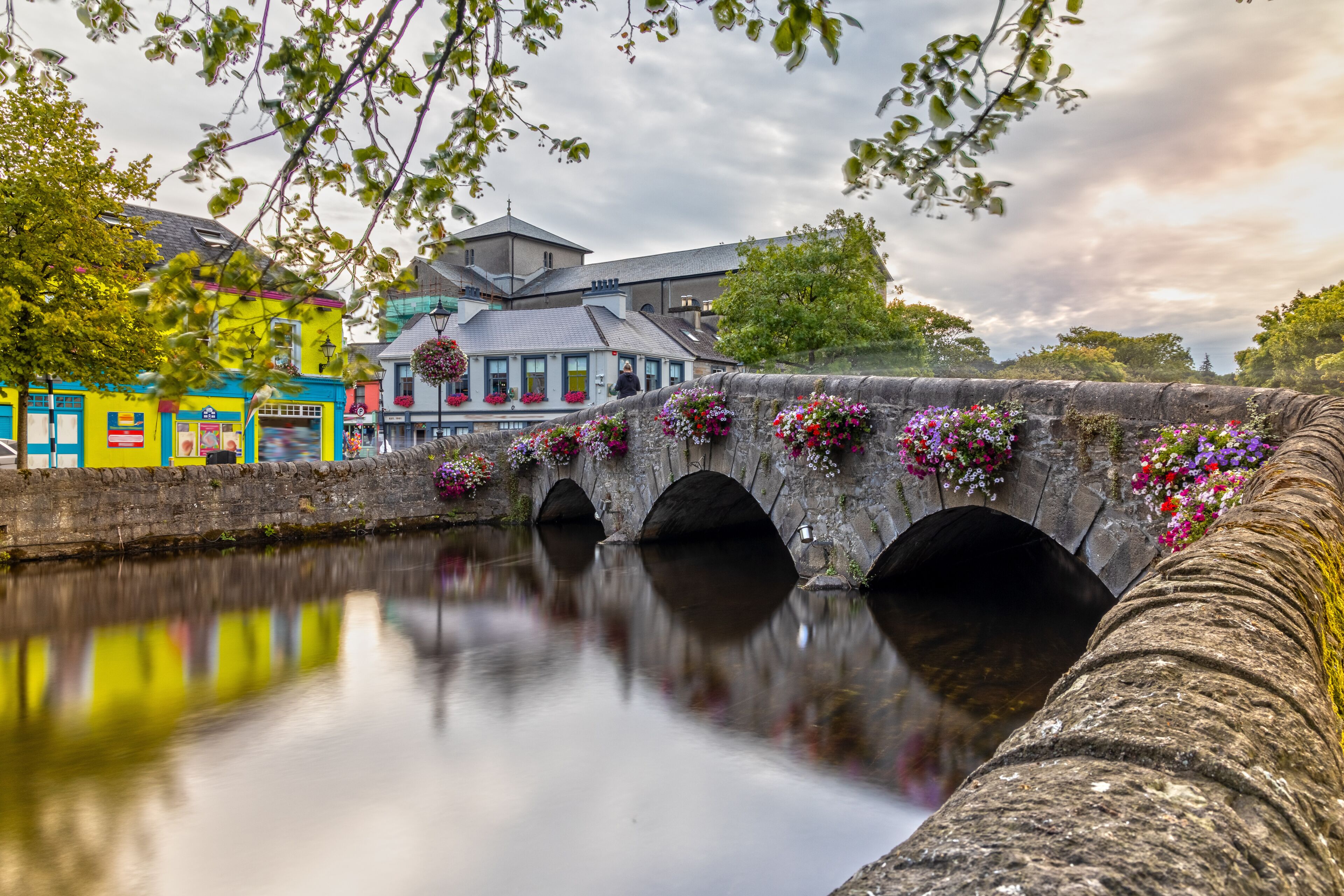 A stone bridge with flower boxes, a calm river, and colorful buildings.