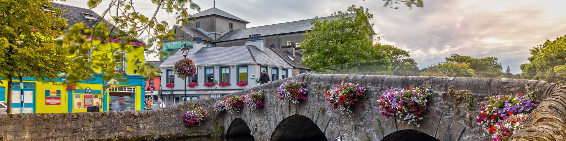 A stone bridge with flower boxes, a calm river, and colorful buildings.