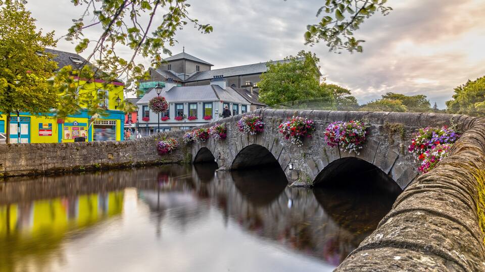 A stone bridge with flower boxes, a calm river, and colorful buildings.