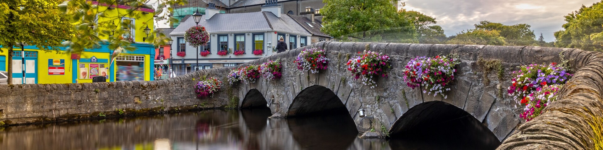 Un puente de piedra con jardineras de flores, un río tranquilo y edificios coloridos.