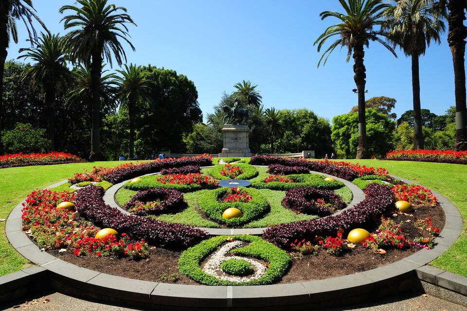 A landscaped garden with a central statue, surrounded by palm trees and a variety of colorful flowers.