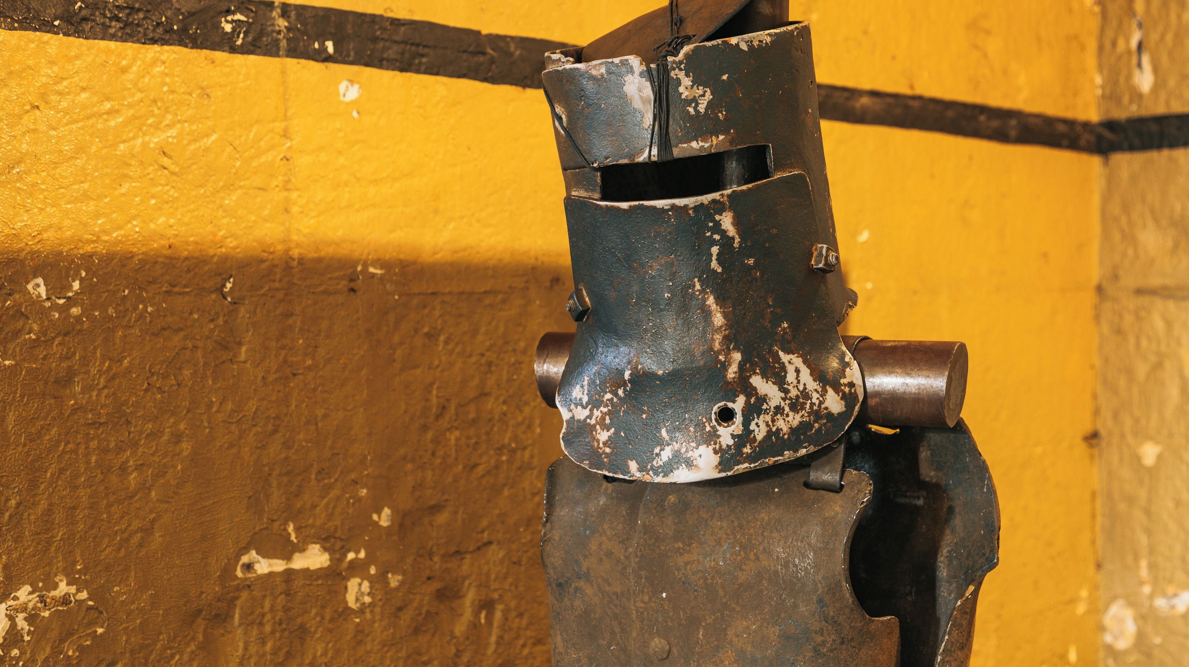 Un objeto de metal oxidado con pintura descascarada frente a una pared amarilla.