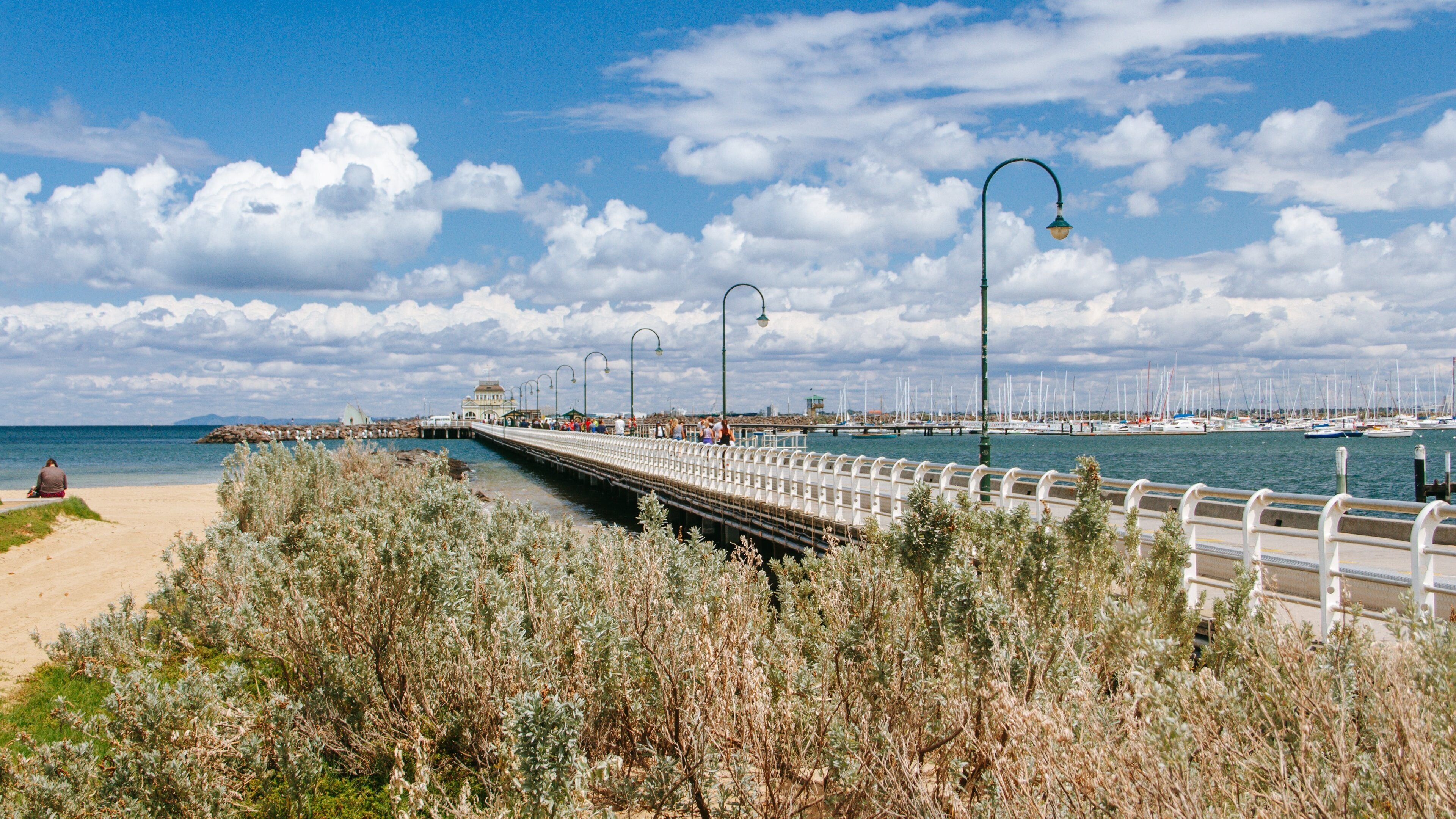 Une promenade en bord de mer, avec un pont, une plage et un port de plaisance où sont amarrés des voiliers.