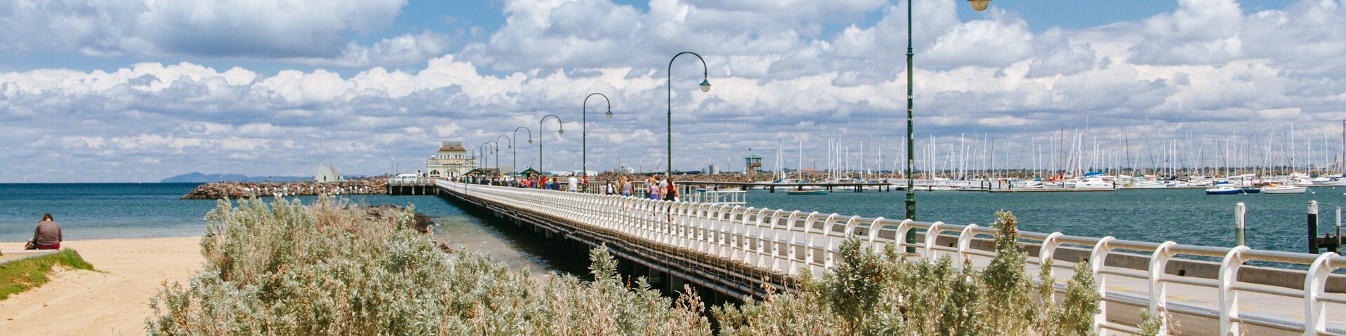 Um calçadão costeiro com uma ponte, uma praia e um porto com veleiros.