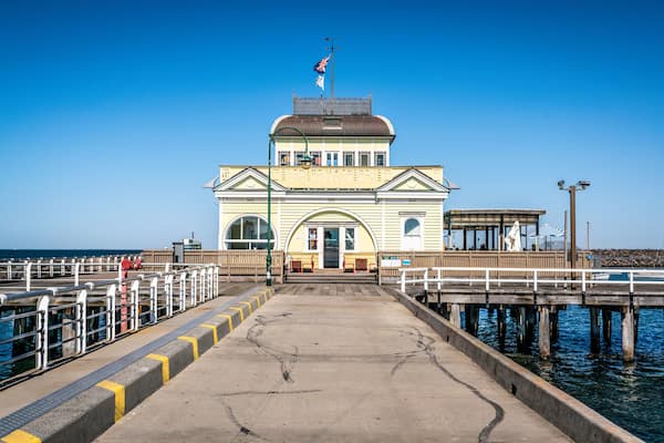 Une jetée menant à un bâtiment jaune surmonté d’un drapeau, une passerelle en bois et un ciel d’un bleu limpide.