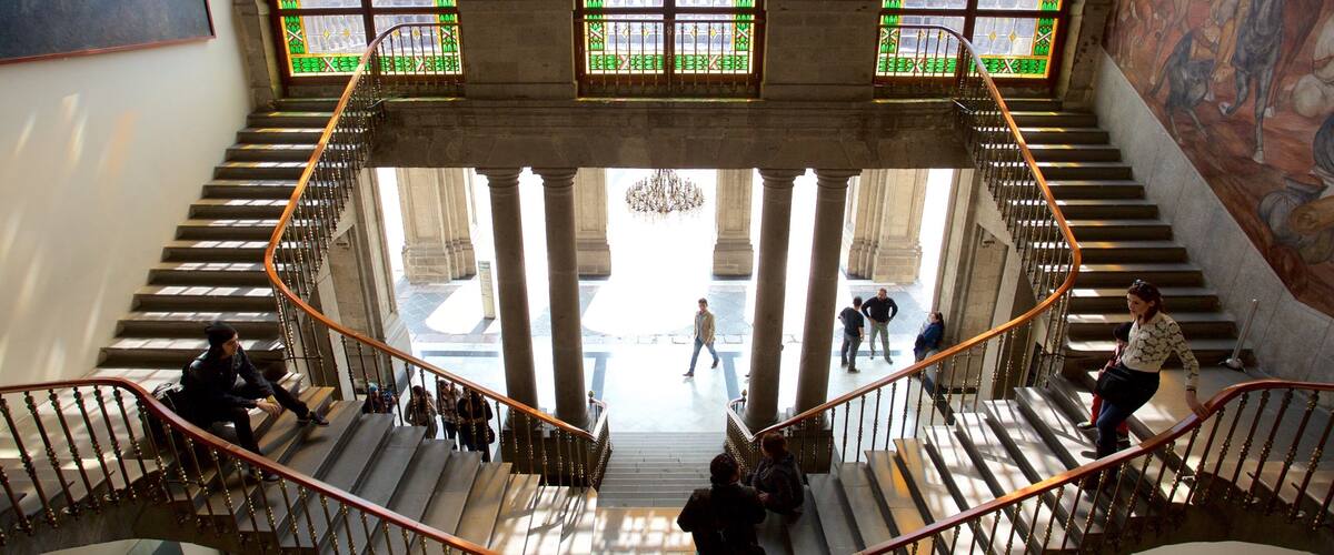 A grand staircase with ornate railings, large windows with green glass panes, and a mural on the wall.