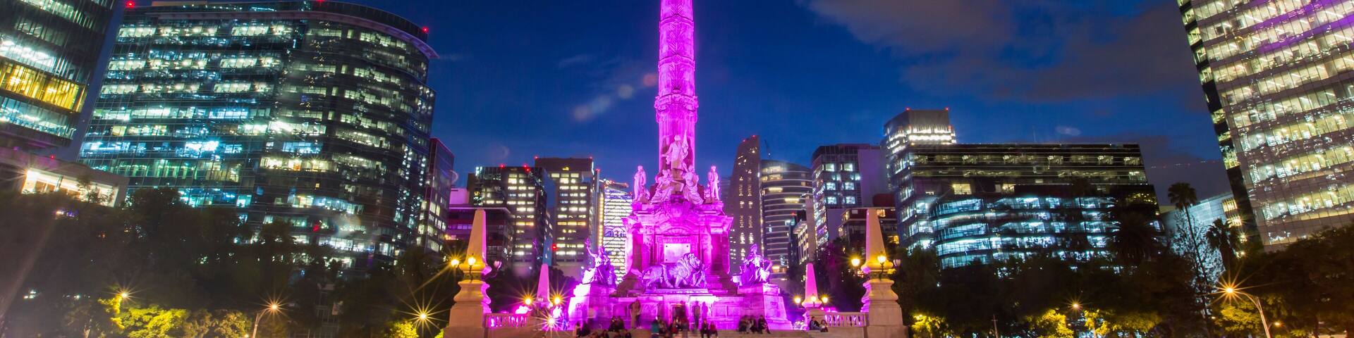 A brightly lit monument with a statue on top, surrounded by modern buildings.