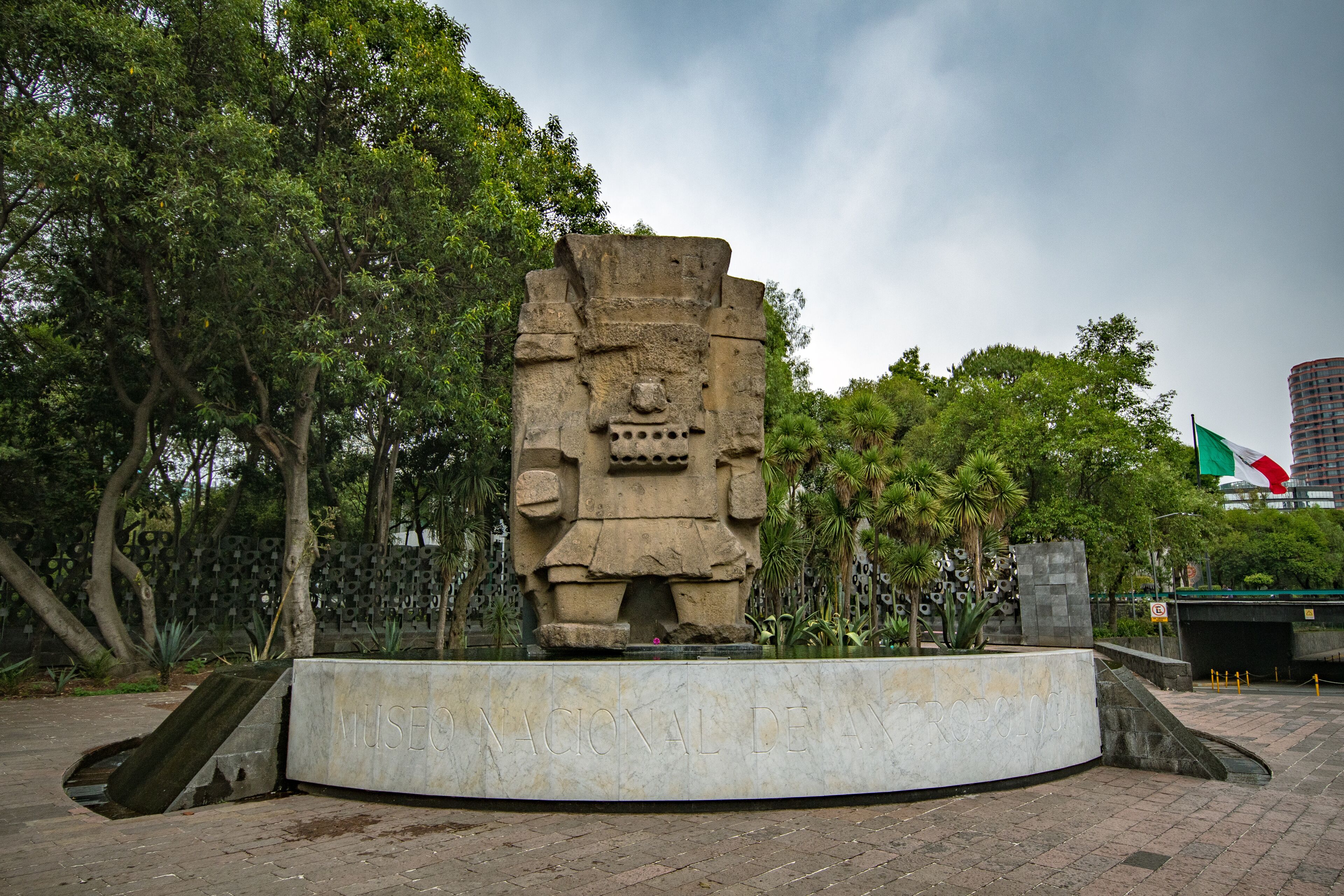 Eine große Steinskulptur in einem Park mit Bäumen und einem modernen Gebäude im Hintergrund.