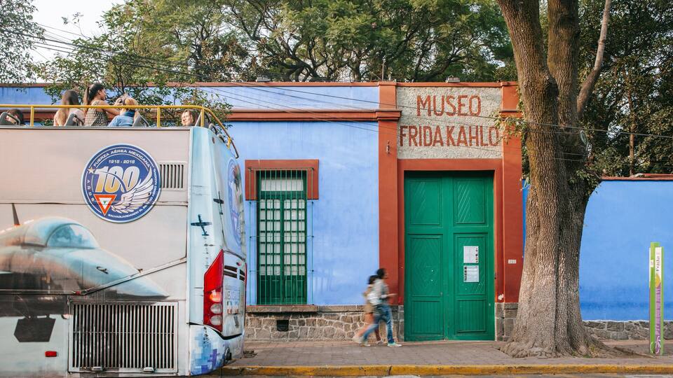 A bus with a blue and white design parked next to a building with a green door labeled "MUSEO FRIDA KAHLO".