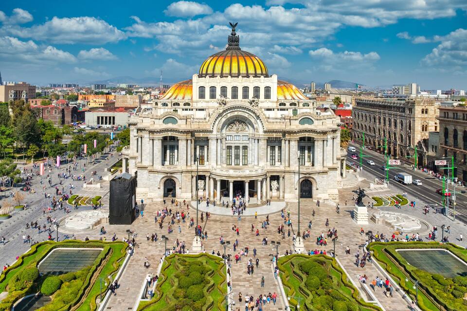 Un gran edificio histórico con una cúpula dorada, rodeado de una plaza animada con gente y vegetación.