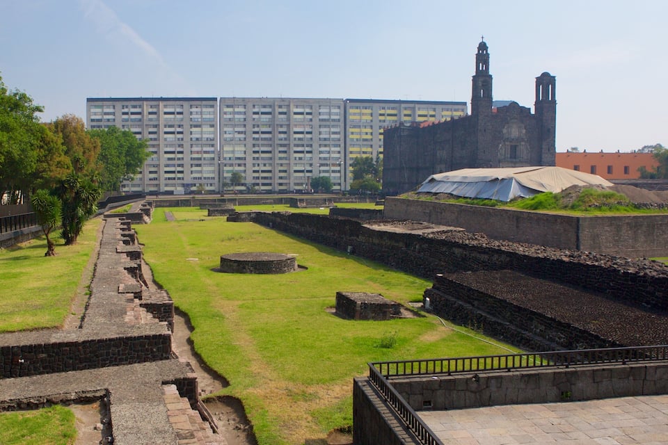 Um sítio arqueológico com ruínas antigas, um edifício grande e uma igreja ao fundo.