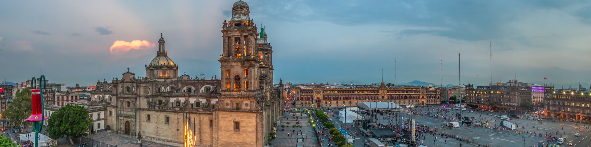 Uma catedral histórica com uma torre de sino imponente, rodeada por uma cidade vibrante com edifícios modernos e uma rua larga.