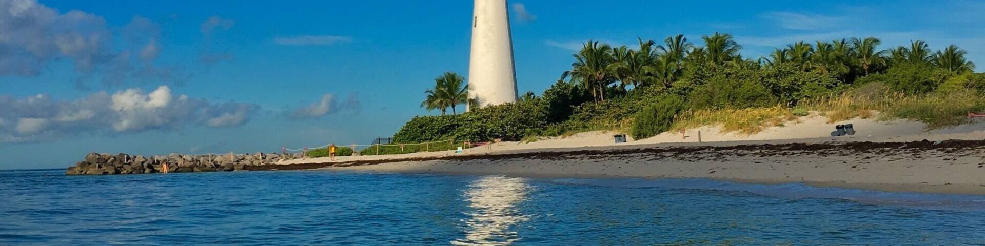 A lighthouse on a sandy beach with palm trees and calm blue waters.