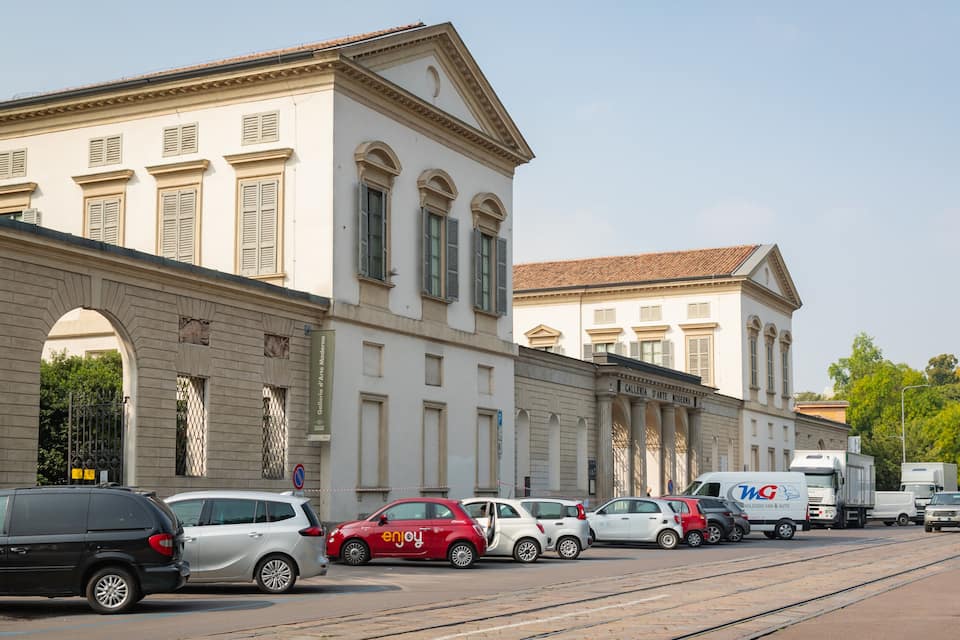 Eine Straße mit parkenden Autos, ein weißes Gebäude mit Säulen und ein Schild mit der Aufschrift 'Galleria d'Arte Moderna'.