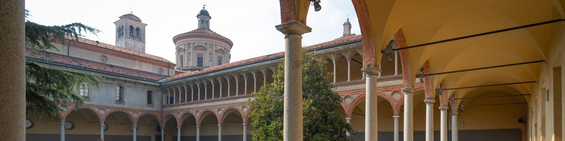 A courtyard with a tiled floor, columns, and a building with a dome and bell tower in the background.