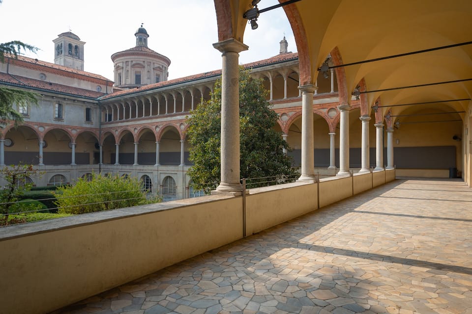 A courtyard with a tiled floor, columns, and a building with a dome and bell tower in the background.