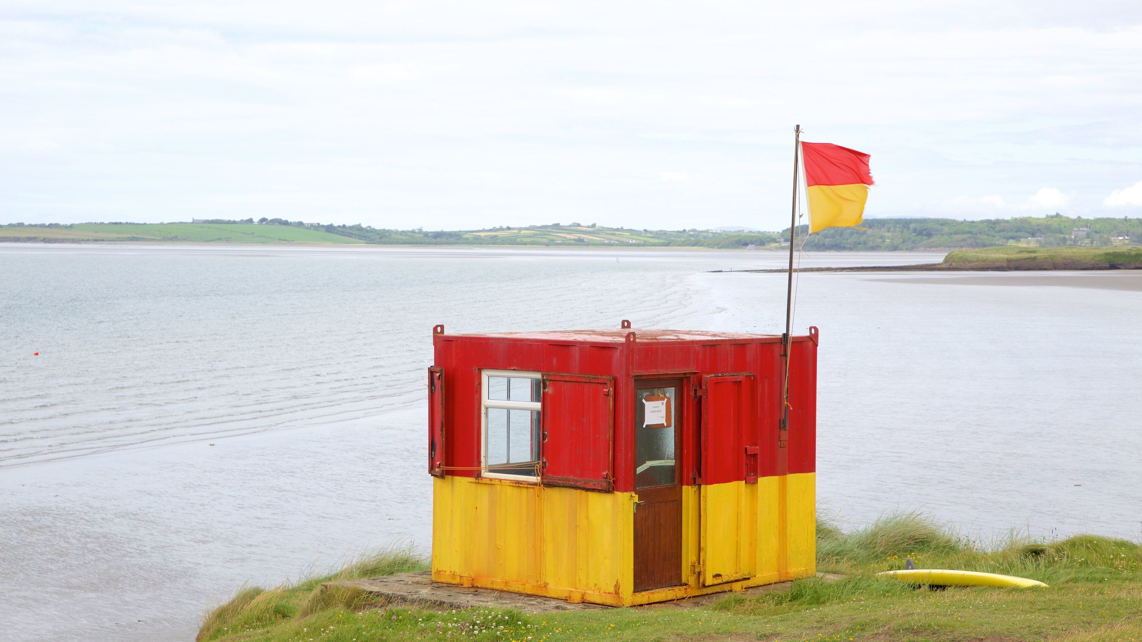 A small red and yellow lifeboat station by a calm lake with a flag on top.