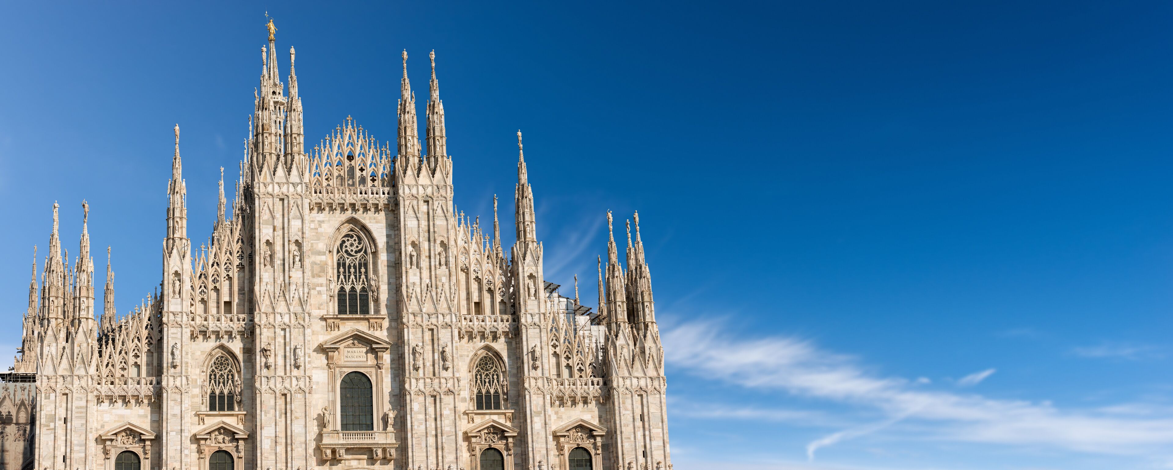 Facade of the Duomo di Milano against a clear blue sky with clouds and copy space. (Milan Cathedral 1418-1577). Church, monument symbol of Lombardy, Italy, Europe.
