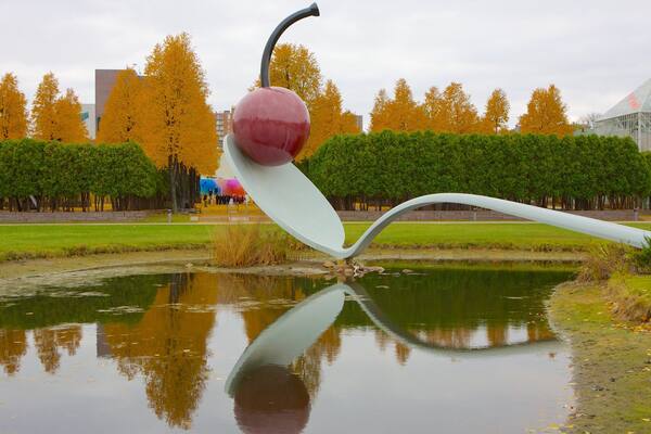 Une sculpture représentant une pomme rouge avec une tige et une feuille blanches, qui se reflète dans un étang, entourée d’arbres aux couleurs automnales et d’un bâtiment moderne.