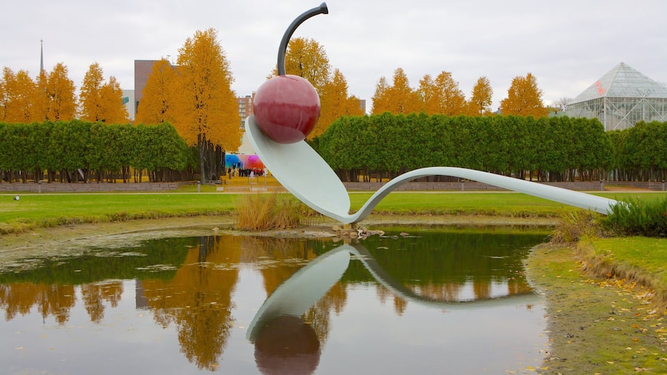A sculpture of a red apple with a white stem and leaf, reflecting in a pond, surrounded by autumn trees and a modern building.