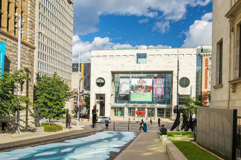 A modern building with a large glass facade, surrounded by trees and a street with pedestrians.